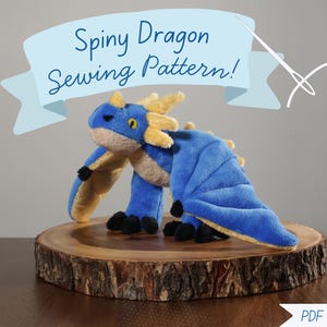 Könnte beinhalten: Ein blau-gelbes Drachen-Plüschtier mit dem Text "Spiny Dragon Sewing Pattern!" auf einem hellblauen Banner. Der Drache hat große Flügel und wird auf einer Holzscheibe präsentiert.
