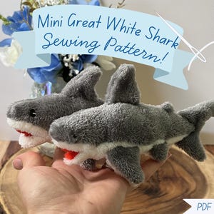 May include: Two small plush great white sharks made of gray and white fabric with black eyes and red mouths. A light blue banner at the top displays the text Mini Great White Shark Sewing Pattern! and a white needle and thread graphic is visible.