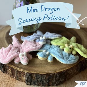 Puede incluir: Varios mini dragones de peluche de diferentes colores, incluyendo rosa, azul y verde, están dispuestos sobre una superficie de madera. La imagen presenta una pancarta con el texto "Mini Dragon Sewing Pattern!" y una ilustración de aguja e hilo. También es visible una opción de descarga en PDF.