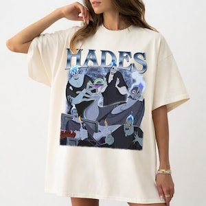 May include: Cream-colored t-shirt featuring a graphic of Hades from Disney's Hercules. The design includes multiple images of Hades with blue flames and the word "HADES" in a blue, metallic font. The shirt has short sleeves and a relaxed fit.