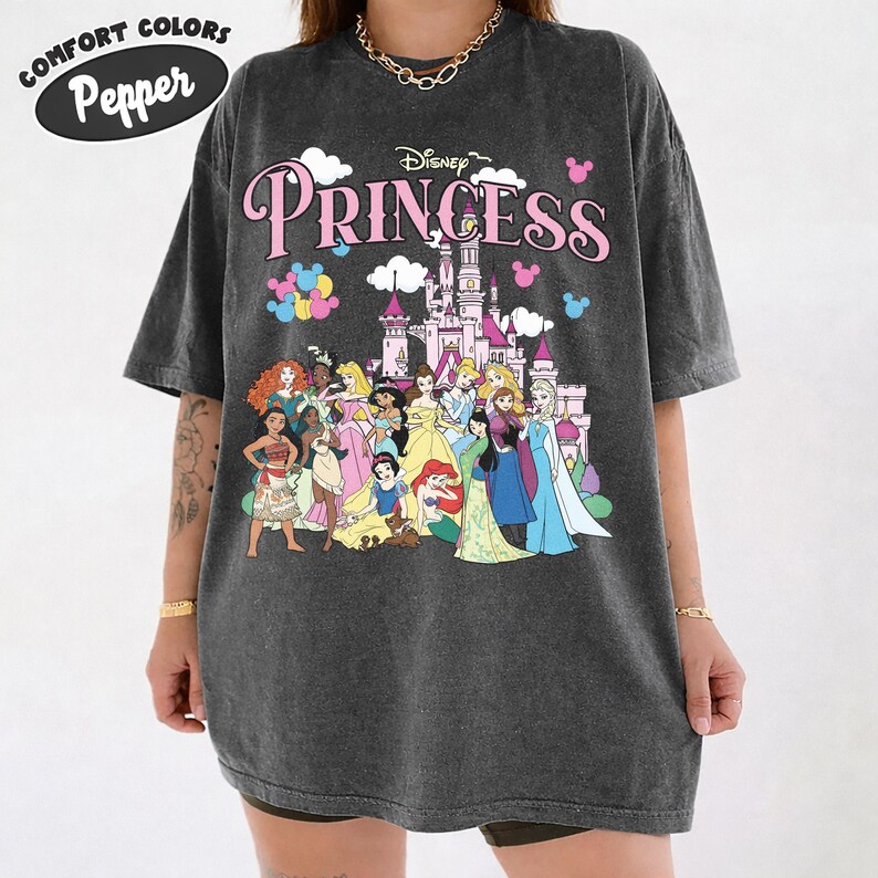 May include: Dark gray Comfort Colors t-shirt featuring a Disney Princess graphic. The design includes the word "Princess" in pink, a castle, and various Disney princesses. The shirt has short sleeves and a relaxed fit.