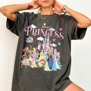 May include: Dark gray t-shirt featuring the word "Princess" in pink script with a Disney logo. The shirt showcases a castle and various Disney princesses in colorful attire. The shirt has short sleeves and a relaxed fit.