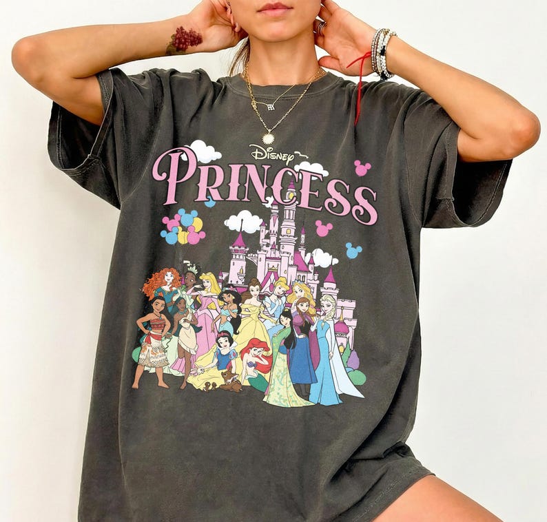May include: Dark gray t-shirt featuring the word "Princess" in pink script with a Disney logo. The shirt showcases a castle and various Disney princesses in colorful attire. The shirt has short sleeves and a relaxed fit.