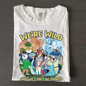 K&ouml;nnte beinhalten: Wei&szlig;es T-Shirt mit einer bunten Grafik mit Zeichentrickfiguren und dem Text "WE'RE WILD AND WE CAN'T BE TAMED". Das Shirt ist gefaltet und hat ein Comfort Colors-Etikett.