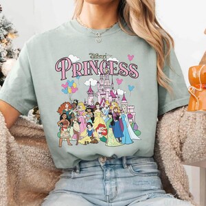 May include: A light green t-shirt featuring the word "Princess" in pink script above a graphic of Disney princesses and a castle. The graphic includes various princesses and colorful balloons. The shirt is a comfortable, casual style.