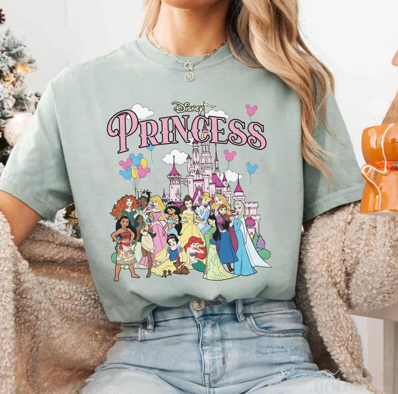 May include: A light green t-shirt featuring the word "Princess" in pink script above a graphic of Disney princesses and a castle. The graphic includes various princesses and colorful balloons. The shirt is a comfortable, casual style.