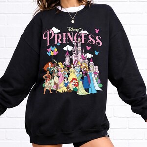 May include: Black sweatshirt featuring a Disney Princess design. The design includes the word "Princess" in pink, a castle, and illustrations of various Disney princesses. The sweatshirt is a crew neck style.