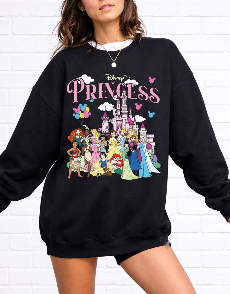 May include: Black sweatshirt featuring a Disney Princess design. The design includes the word "Princess" in pink, a castle, and illustrations of various Disney princesses. The sweatshirt is a crew neck style.