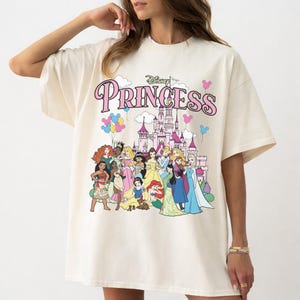 May include: Cream-colored t-shirt featuring the word "Princess" in pink script above a castle illustration and a group of cartoon princesses. The shirt has short sleeves and a relaxed fit.