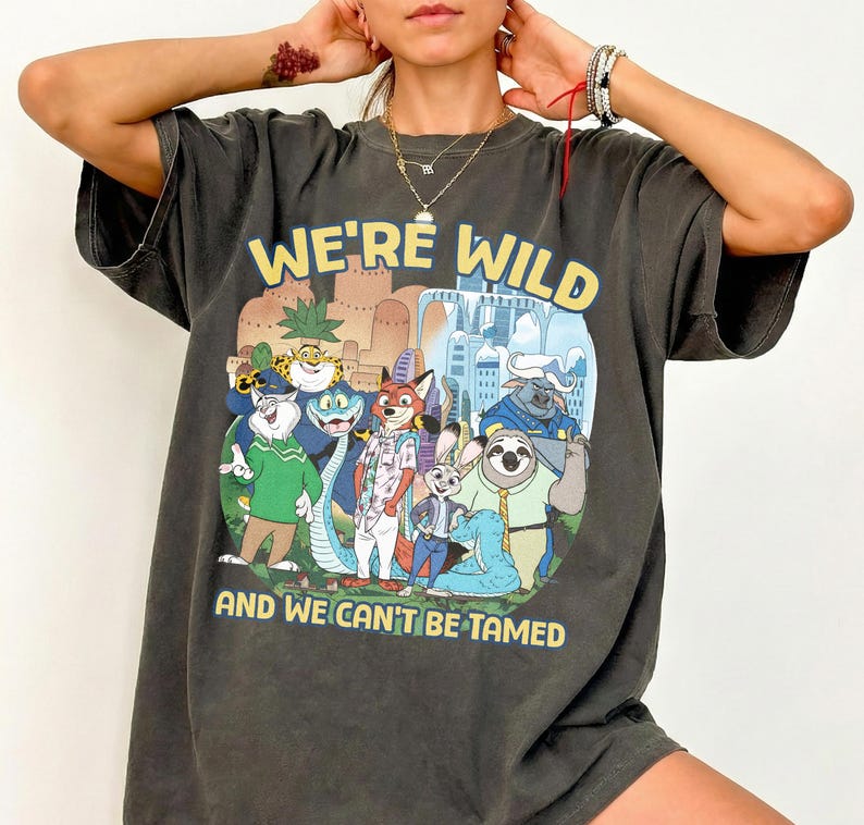 K&ouml;nnte beinhalten: Dunkelgraues T-Shirt mit einem farbenfrohen Grafikdruck von Zeichentrickfiguren und dem gelben Text "WE'RE WILD AND WE CAN'T BE TAMED". Das Shirt hat kurze &Auml;rmel und eine lockere Passform.