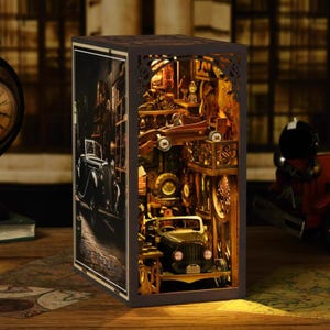 May include: A wooden, illuminated diorama depicting a vintage car repair shop. The detailed scene includes a classic car, tools, and spare parts, all within a dark brown, open-sided box. The left side features a black and white street scene.