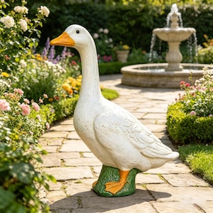 Może przedstawiać: Duży, biały posąg gęsi z pomarańczowym dziobem i stopami stoi na zielonej podstawie. Posąg znajduje się w ogrodzie ze ścieżką z kamienia, kwiatami i fontanną w tle. Ozdoba do ogrodu.