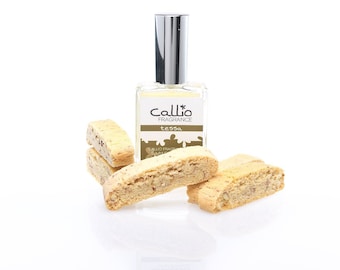 May include: A clear glass bottle of Callio Fragrance Tessa perfume with a silver cap. The bottle is sitting on a white surface with several biscotti cookies.