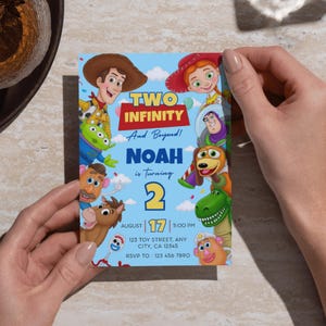 May include: A rectangular birthday invitation card with a light blue background featuring cartoon characters including a cowboy, cowgirl, space ranger, green alien, dinosaur, and potato figures. Text reads: TWO INFINITY And Beyond! NOAH is turning 2, AUGUST 17, 5:00 PM, 123 TOY STREET, ANY CITY, CA 12345, RSVP TO: 123 456 7890.