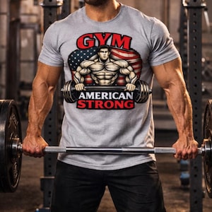 Peut inclure: T-shirt gris avec un motif d'haltérophile, le drapeau américain et les mots "GYM" et "AMERICAN STRONG". Le t-shirt est porté avec un short noir et une casquette noire. L'image est prise dans une salle de sport.