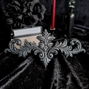 May include: A decorative wall appliqué featuring an intricate, symmetrical acanthus leaf scrollwork design. The piece is finished in a metallic silver-grey tone with dark shading in the recessed areas to highlight the detailed, carved-style relief.