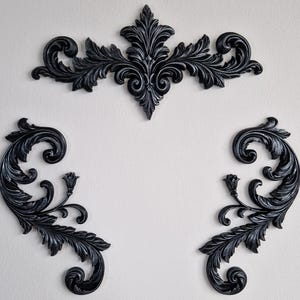 May include: Three black wall decor pieces featuring intricate carved acanthus leaf scrollwork. One large central horizontal piece with symmetrical foliage patterns and two smaller matching side pieces with curved stems and floral accents.