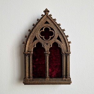 May include: A decorative wall-mounted display frame featuring a bronze-coloured finish with a pointed gothic arch design. The structure includes intricate architectural detailing, a quatrefoil cutout at the top, and a deep red velvet backing.
