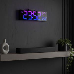 Może przedstawiać: Czarny cyfrowy zegar wyświetla godzinę w kolorze niebieskim i różowym. Poniżej czarny soundbar i biały wazon na pływającej półce. Roślina doniczkowa z zielonymi liśćmi dodaje odrobinę natury do minimalistycznego wystroju.