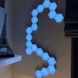 May include: A wall-mounted light installation composed of blue, hexagonal LED panels. The panels are arranged in a flowing, curved design. The lights emit a bright blue glow. A miniature Eiffel Tower model is visible on a shelf in the background.