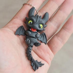 May include: A black dragon keychain with a smiling expression, yellow eyes, and a red tongue. The dragon has small wings and a segmented tail. The keychain is attached to a silver chain.