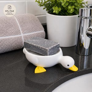 May include: A white ceramic duck-shaped soap dish with a gray bar of soap. The duck has yellow feet and a yellow beak. A gray towel and a small potted plant are in the background. The soap dish is on a black countertop.