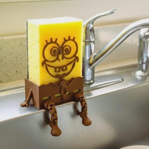 May include: A yellow sponge with a brown cartoon face and legs, sitting on the edge of a stainless steel sink. The sponge is held in place by a brown holder. A chrome faucet is in the background.