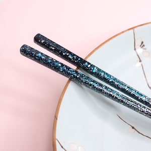 May include: A pair of chopsticks featuring a dark blue and black base with a shimmering, multi-colored glitter finish. The upper sections are cylindrical and textured with reflective particles, while the lower tips taper to a smooth, solid brown finish.