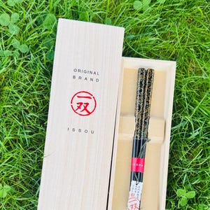 May include: A pair of black chopsticks with gold-flecked handles inside a light-colored wooden box. The lid features the text ORIGINAL BRAND ISSOU and a red circular logo. A red band with the text ISSOU wraps around the chopsticks.