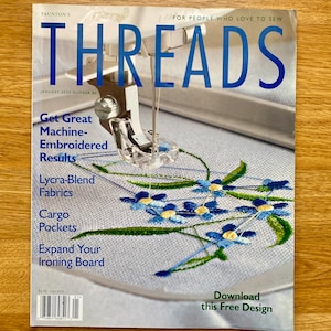Puede incluir: Una portada de revista para la revista Threads, que presenta un primer plano de una aguja de máquina de coser y un hilo que borda un diseño de flores azules sobre una tela blanca. El texto de la portada de la revista dice "Obtenga excelentes resultados de bordado a máquina", "Tejidos de mezcla de Lycra", "Bolsos de carga", "Amplíe su tabla de planchado" y "Descargue este diseño gratuito".