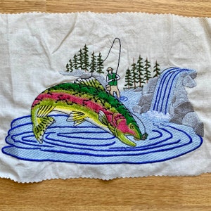 Puede incluir: Diseño bordado de una trucha arcoíris saltando fuera del agua, con un pescador de pie en la orilla. La escena incluye una cascada y pinos.