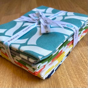 May include: A stack of colorful fabric squares tied with a white ribbon. The fabric features a teal and white floral pattern. The ribbon is printed with the words "Cloud Fabrics". The edges of the fabric squares are visible, showing a variety of colors.