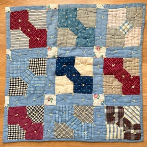 Puede incluir: Una colcha de retazos azul con una variedad de patrones, incluyendo cuadros, vichy y lunares. La colcha está compuesta por nueve cuadrados, cada uno con un patrón diferente. La colcha está atada con cuerda blanca.