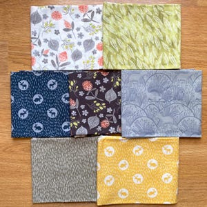 K&ouml;nnte beinhalten: Eine Auswahl gefalteter Stoffquadrate mit verschiedenen von der Natur inspirierten Mustern. Designs umfassen Blumen-, Blatt- und Waldtiermotive in Farben wie Wei&szlig;, Gr&uuml;n, Marine, Braun, Grau und Gelb. Die Stoffe sind auf einer Holzoberfl&auml;che angeordnet.