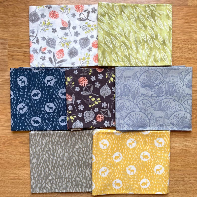 K&ouml;nnte beinhalten: Eine Auswahl gefalteter Stoffquadrate mit verschiedenen von der Natur inspirierten Mustern. Designs umfassen Blumen-, Blatt- und Waldtiermotive in Farben wie Wei&szlig;, Gr&uuml;n, Marine, Braun, Grau und Gelb. Die Stoffe sind auf einer Holzoberfl&auml;che angeordnet.