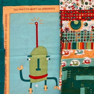 May include: A blue fabric panel with the text "You make my heart go whirbrrrr" printed on it. The panel features a cartoon robot with a red antenna, green body, and red legs. The robot has a large, green head with a red mouth and black eyes. The panel is surrounded by a variety of colourful fabric scraps with geometric patterns.