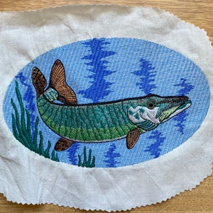 Puede incluir: Diseño bordado de un pez muskellunge verde y marrón nadando en agua azul con juncos verdes. El pez está en una forma ovalada.