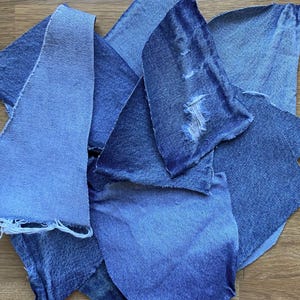 May include: An assortment of denim fabric scraps in shades of blue, from light to dark. Some pieces show signs of wear and tear, with frayed edges and distressed areas. The pieces are arranged in a haphazard manner, overlapping each other.