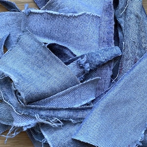 Puede incluir: Primer plano de varios trozos de tela vaquera azul claro con bordes deshilachados. Las piezas de mezclilla están dispuestas en un patrón superpuesto en capas, mostrando la textura y el tejido del material. La tela parece nueva.