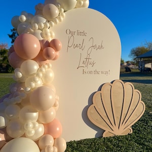 May include: A cream-colored, semi-circular sign with the text "Our little Pearl Jerah Lattus Is on the way!" is decorated with a balloon garland in shades of white and pink. A wooden seashell cutout is placed at the bottom.
