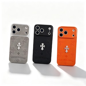 Puede incluir: Tres fundas para smartphone en gris, negro y naranja. Cada funda presenta un emblema de cruz plateado en el centro, patrones de cruces en relieve en la parte posterior y el texto CHROME HEARTS impreso cerca del borde inferior de la funda.