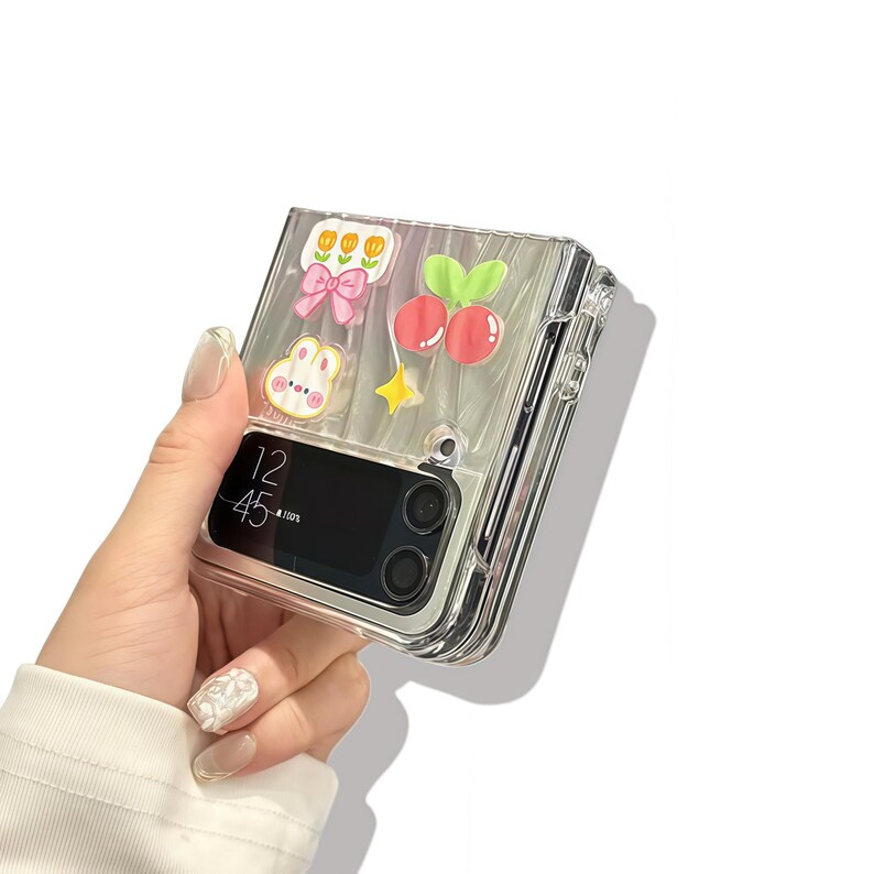 Peut inclure: Coque de t&eacute;l&eacute;phone transparente orn&eacute;e de d&eacute;corations color&eacute;es de style dessin anim&eacute;. La coque pr&eacute;sente un n&oelig;ud rose, des cerises, une &eacute;toile et un lapin. L'&eacute;cran du t&eacute;l&eacute;phone affiche l'heure 12h45. Con&ccedil;u pour un t&eacute;l&eacute;phone pliable.