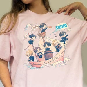 May include: A light pink t-shirt featuring a cartoon illustration of a ship with seven animated characters. The word "SWIM" is in blue, and the ship is sailing on blue waves. The shirt is made of a soft material.