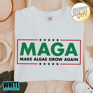 May include: A white short-sleeved t-shirt featuring a graphic design with a red rectangular border. Inside the border, the green text MAGA is displayed above the black text MAKE ALGAE GROW AGAIN, flanked by five black stars above and below.