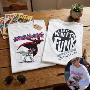 May include: Two white t-shirts with graphic designs. One shirt features the word "PARLIAMENT" in blue and red, with a cartoon figure. The other shirt says "WE WANT THE FUNK" with "GEORGE CLINTON" below. Sunglasses and vintage magazines are also visible.