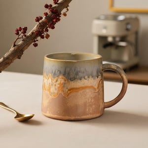 May include: A ceramic mug with a reactive glaze featuring a gradient of cool grey at the rim, a textured amber band in the middle, and a warm tan base. A small gold-toned metal spoon rests beside the mug, and a branch with red berries hangs above.