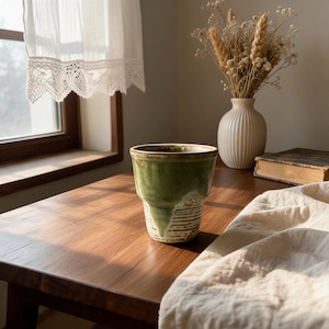 May include: A ceramic cup featuring a two-tone design with a smooth, glossy olive green glaze on the upper portion and a textured, cream-coloured base with horizontal ridges. The cup has a wide, open rim and a tapered body shape.