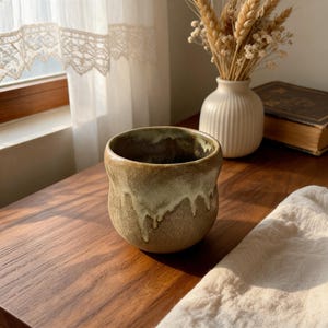 Puede incluir: Una taza de cerámica redondeada con una base texturizada de color marrón tierra y un esmalte de color crema que gotea desde el borde. El recipiente tiene una boca ancha y una silueta curva y suave, mostrando un aspecto artesanal.