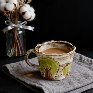 May include: A ceramic mug with a wavy rim and a handle, featuring a raised green tree design on a cream-colored base with brown speckles. The mug is filled with a light brown liquid and sits on a gray fabric napkin next to a glass vase.