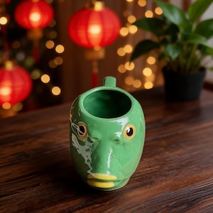 Może przedstawiać: Zielony ceramiczny kubek w kształcie głowy ryby z uchwytem na górze. Kubek posiada dwoje ciemnych oczu, teksturowaną powierzchnię i żółte usta. Projekt jest uformowany tak, aby przypominał rysy twarzy ryby, z błyszczącym wykończeniem na całej powierzchni.